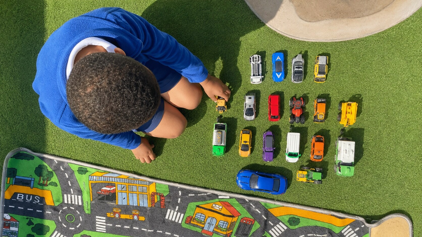 Child carefully arranging toy cars beside a road mat during outdoor play