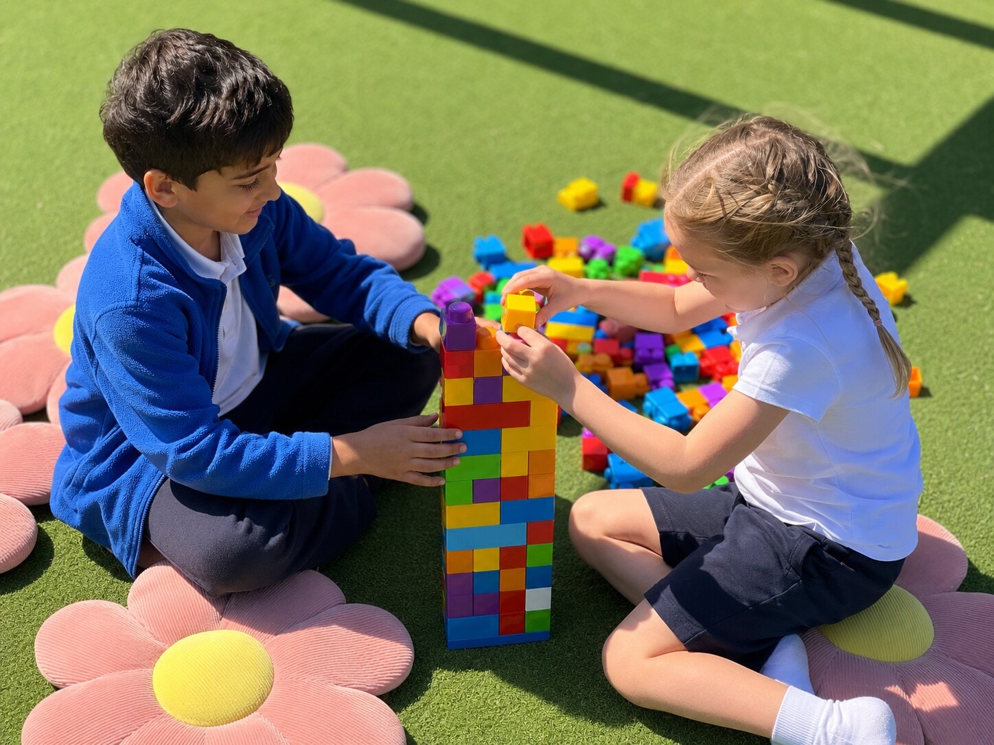 Two children collaborating to build a tall tower with colourful blocks on artificial turf