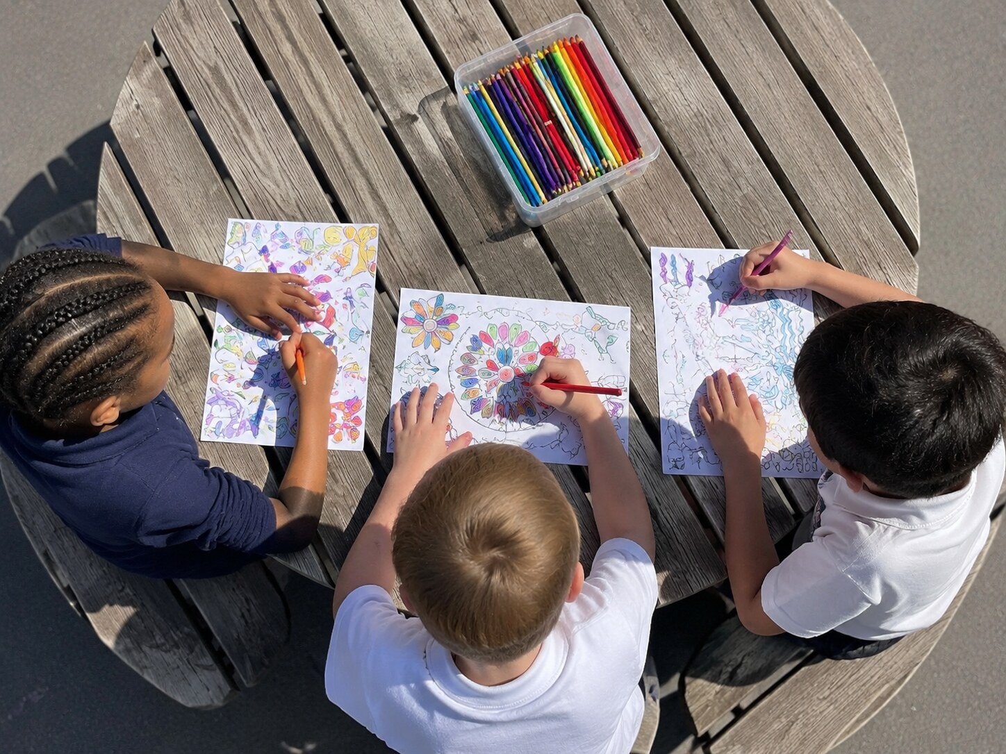 Four children colouring artwork together at an outdoor table with coloured pencils
