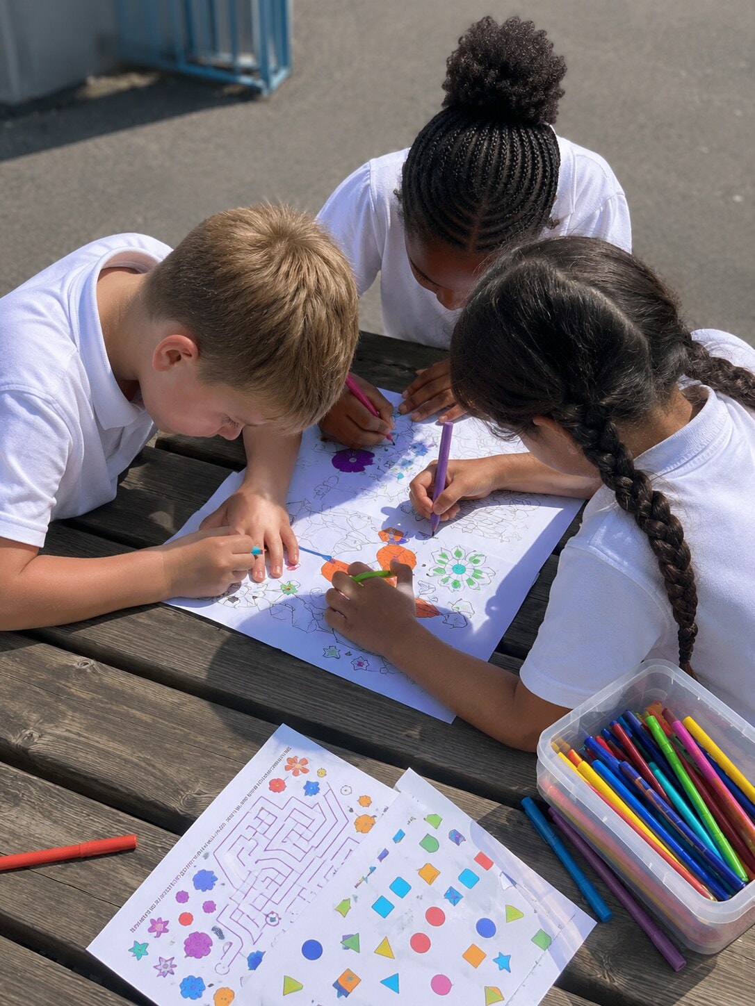 Three children colouring and drawing together at an outdoor table with felt-tip pens